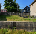 photo showing vacant 23 Water Street lot with grass and weeds growing behind a fence. 