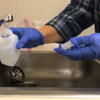 A silver sink with a woman wearing rubber gloves to fill up a plastic bottle while gathering a water sample from the running tap