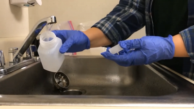 A silver sink with a woman wearing rubber gloves to fill up a plastic bottle while gathering a water sample from the running tap