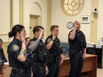 An image showing four new police officers in uniform holding their right hand up as they are sworn in at a Select Board meeting.